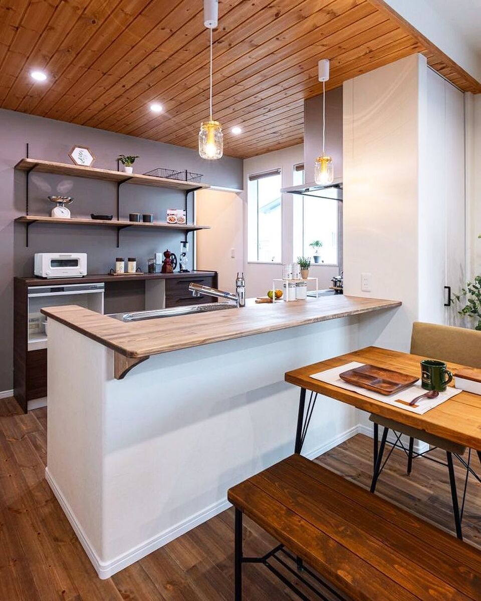 天井と床の無垢材に合わせた木製のカウンタートップが白い壁に映えるナチュラルテイストな対面キッチン｡グレーのアクセントクロスと可愛いペンダントライトでますますおしゃれなキッチンに！