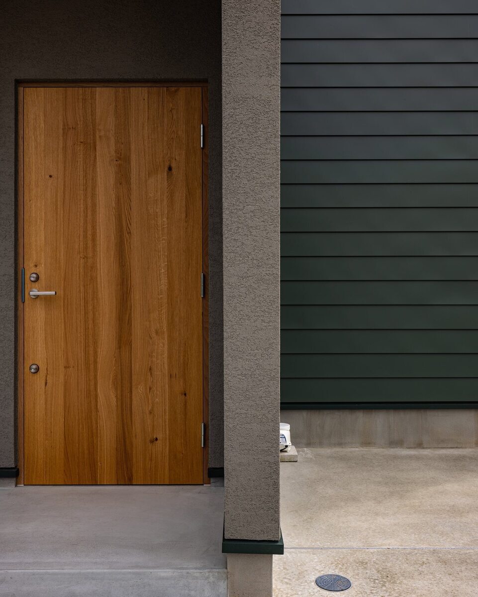 モスグリーンのガルバリウムと、ブラウンの塗り壁×木目の玄関ドアがシックに調和したおしゃれな外観