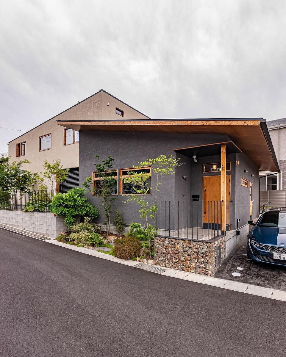 グレーの外壁がシックな平屋建てと、ベージュの外壁がナチュラルな２階建てを組み合わせたインパクトのあるデザインのモダンな家。植栽の美しい緑が二棟の建物を緩やかにつなげています。